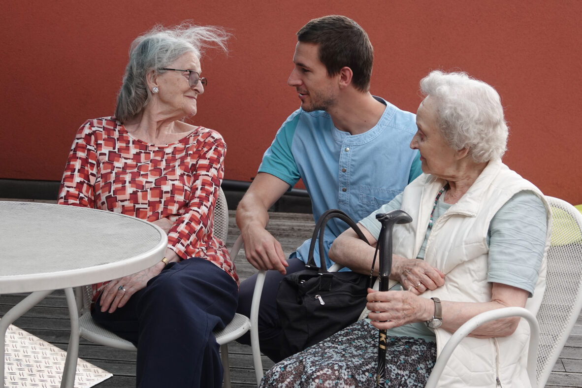 ARC 3 Twee bewoners van het neuropsychologisch woonzorgcentrum Arcus in Brussel zitten samen op het terras, in een gezellig gesprek met een medewerker die naast hen gebukt is, zodat ze elkaar op ooghoogte kunnen aankijken.