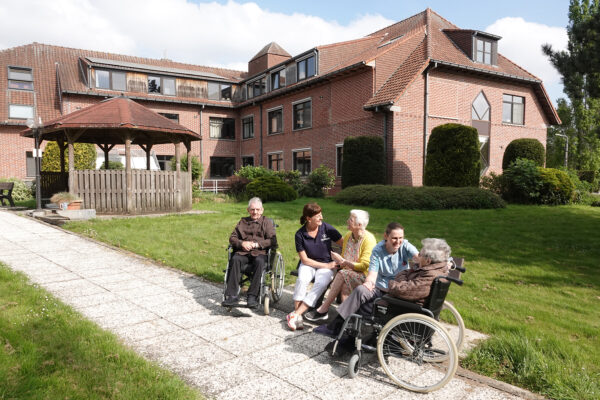 De tuin van woonzorgcentrum De Vlaamse Ardennen, waar drie bewoners samen met twee medewerkers op een bankje zitten. Op de achtergrond is het bakstenen gebouw duidelijk zichtbaar. Het pad in de tuin is goed begaanbaar, waardoor de tuin voor iedereen toegankelijk is, ook voor bewoners die een rolstoel gebruiken.