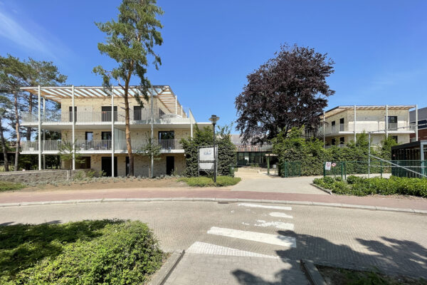 Straatzicht van woonzorgcentrum Angelahof, waarbij het pad naar de ingang duidelijk zichtbaar is. Het gebouw is ook te zien, met op elke verdieping een groot omringend terras.