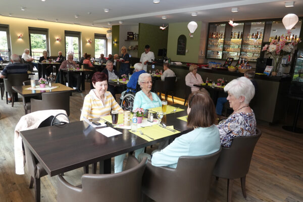 De brasserie van woonzorgcentrum Aurora, waar enkele bewoners zitten te wachten op hun bestelling. Ze hebben hun eten besteld en wachten geduldig totdat het wordt geserveerd. Op de achtergrond is een van de koks aanwezig, die controleert of alles goed verloopt en of de bewoners tevreden zijn.
