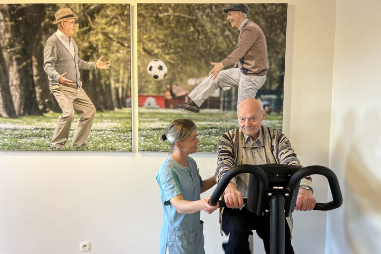 BOS 1 De kinesitherapieruimte in woonzorgcentrum Berckenbosch in Heusden. Een bewoner voert fietsoefeningen uit op een hometrainer, terwijl een medewerkster hem ondersteunt en aanmoedigt. Op de achterwand hangen twee grote foto's die samen een geheel vormen, waarop twee oudere mannen te zien zijn. Eén man schopt een voetbal naar de andere.