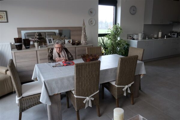 Een bewoner van een serviceflat in woonzorgcampus Berkenhof zit in haar eetkamer aan een tafel die plaats biedt aan zes personen en leest een tijdschrift. Achter haar staat een commode met familiefoto's en decoraties. Aan de muur hangen twee borden en een klok.