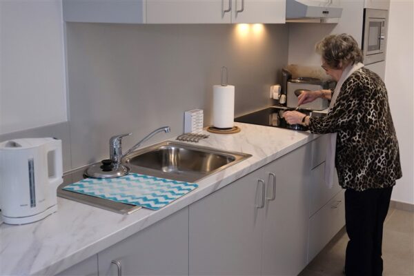 De keuken van een serviceflat in woonzorgcampus Berkenhof. De bewoner roert in een pot op de kookplaat. Op het keukenblad staan een pillendoos, een waterkoker en een rol keukenpapier. De keukenkasten zijn lichtgrijs en hebben metalen handvatten.
