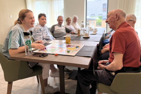 Negen bewoners zitten samen aan een lange tafel en genieten van een drankje. Een logistieke medewerkster van woonzorgcentrum Bloemenhof is bij hen en speelt een bordspel met een van de bewoners, terwijl de anderen toekijken.
