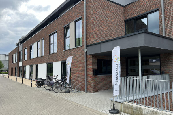 De ingang van woonzorgcentrum Bloemenhof, gemarkeerd door verticale witte vlaggen met de naam en het logo van het woonzorgcentrum. Bij de ingang staat een fietsenrek, waar meerdere fietsen en een motor geparkeerd staan.