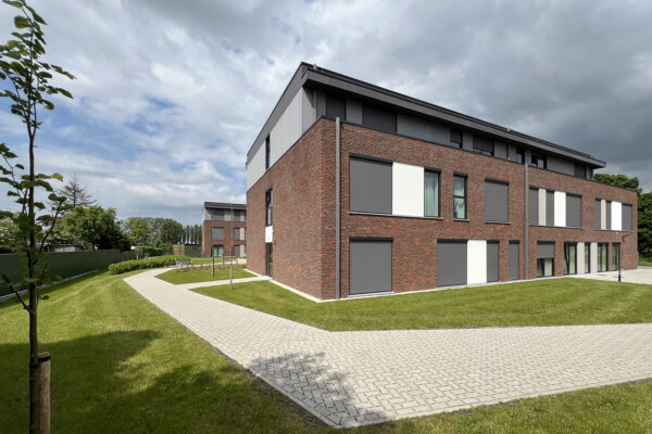 De achtergevel van woonzorgcentrum Bloemenhof. De meeste ramen zijn bedekt om het zonlicht te weren. De begane grond en eerste verdieping hebben gevels van baksteen, terwijl de tweede verdieping kleiner is en gevels heeft die bekleed zijn met grijze platen.