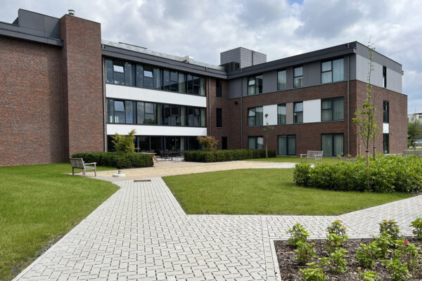 De buitenzijde van het gebouw van woonzorgcentrum Bloemenhof. De tuin is voorzien van verschillende stenen paden en twee houten banken. Het gras is goed onderhouden en zorgvuldig gemaaid. Het gebouw heeft gevels van baksteen, met witte accenten.