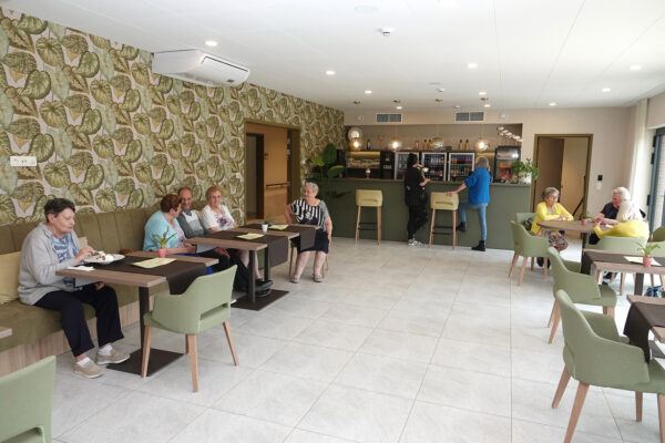 Een overzicht van het Grand Café van woonzorgcentrum Bloemenhof. Het café heeft een groene bar met twee barstoelen waar bezoekers iets kunnen bestellen. Twee groepen bewoners zitten aan verschillende tafels, terwijl een andere bewoner alleen aan een tafel zit en geniet van een dessert.