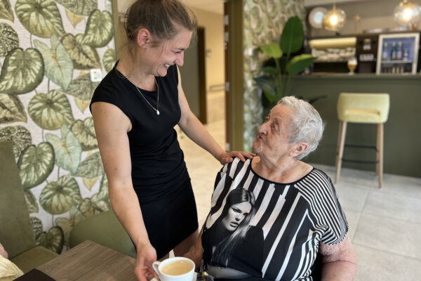 De directrice van woonzorgcentrum Bloemenhof brengt een kopje koffie naar een bewoner die in het Grand Café zit. De twee kijken elkaar aan, waarbij de directrice haar hand op de schouder van de bewoner legt en een brede glimlach toont.