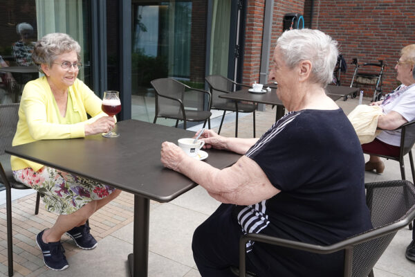 Twee bewoners zitten samen op het terras van het Grand Café van woonzorgcentrum Bloemenhof. De één drinkt een bier, de ander een kopje koffie. Aan een andere tafel zitten twee andere bewoners, die ook samen iets aan het drinken zijn.