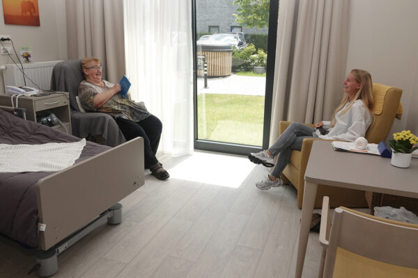 Een dochter bezoekt haar moeder in woonzorgcentrum Bloemenhof. De dochter zit in een gele zetel tegenover haar moeder, die in een andere zetel zit, bedekt met een grijze deken. De moeder houdt een blauw boek in haar handen. De kamer heeft directe toegang tot de tuin via een grote schuifdeur.