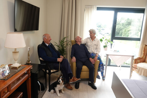 Drie personen en een klein hondje zitten samen in een kamer van woonzorgcentrum Bloemenhof. Eén van hen zit op een zwarte stoel, terwijl de andere man op een gele zetel zit en met een brede glimlach naar een vrouw kijkt die op de leuning van de zetel zit. De kamer heeft een groot raam dat veel licht binnenlaat. Er staat in de kamer een kleine zwarte koelkast, een andere fauteuil, een televisie aan de muur, een bed en een bijzettafel met een sierlijke lamp en een familiefoto.