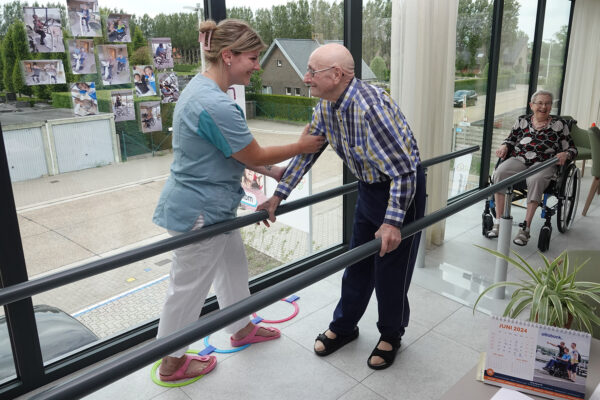 De kinesitherapieruimte van woonzorgcentrum Bloemenhof. Met de hulp van een verpleegster voert een bewoner stapoefeningen uit, terwijl hij zich aan leuningen vasthoudt. Een vrouwelijke bewoner zit op haar rolstoel te wachten op haar beurt. Op het raam hangen foto's van bewoners en medewerkers die verschillende oefeningen uitvoeren. Op de voorgrond is een kalender te zien, geopend op de maand juni van 2024.