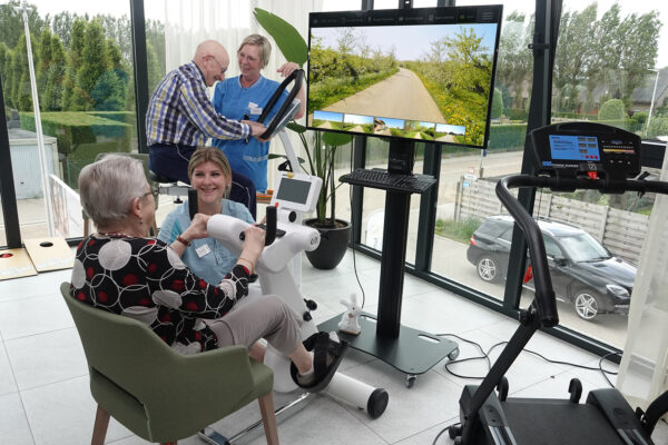 In de kinéruimte van woonzorgcentrum Bloemenhof voeren twee bewoners fietsoefeningen uit, beide met de hulp van een verpleegster. De vrouw gebruikt een hometrainer die verbonden is met een televisie, waarop een pad wordt weergegeven, waardoor het lijkt alsof ze buiten fietst. De mannelijke bewoner gebruikt een klassieke hometrainer. In de ruimte is ook een loopband beschikbaar.