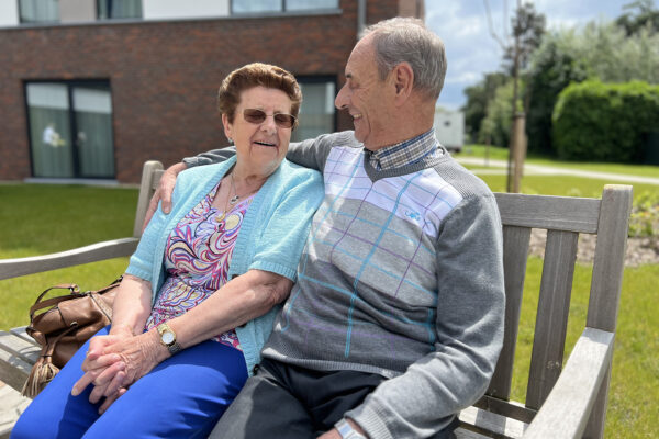 Twee bewoners van woonzorgcentrum Bloemenhof zitten samen op een houten bank in de tuin. De mannelijke bewoner heeft zijn arm om de vrouwelijke bewoner geslagen. Beide hebben hun ogen gesloten, terwijl de vrouw een zonnebril draagt. Naast haar staat haar bruine tas.