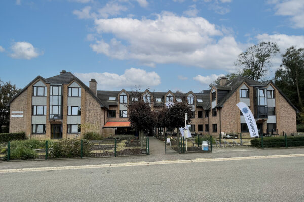 Straatzicht op het symmetrische gebouw van woonzorgcentrum Boneput in Bree. Vlak voor de ingang bevindt zich een bushalte. Om bij de ingang te komen, loopt men over een pad dat aan beide zijden omringd is door bomen.