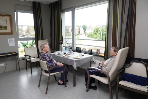 Een dubbele kamer waar een man en zijn vrouw samenwonen in woonzorgcentrum Damiaan. Ze zitten beiden aan een tafel te rusten na hun maaltijd. De kamer is ingericht met vijf bijpassende stoelen, en tegen de muur staat een elektrische piano. Op de vensterbank liggen verschillende familiefoto's.