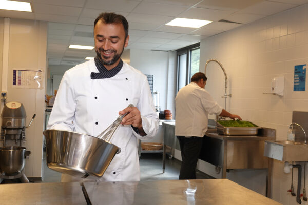 In de keuken van woonzorgcentrum Sporenpark bereidt de chef-kok een taart. Op de achtergrond is een andere kok druk bezig met het wassen van de groenten die later in de maaltijd zullen worden verwerkt.