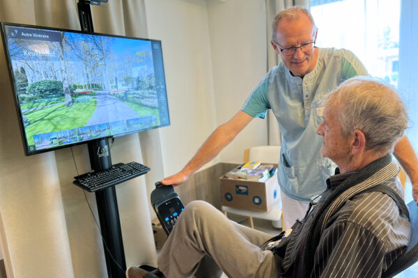 Les Charmilles - Salle de kiné une personne âgée est sur un vélo d'appartement devant une tv - maison de repos - Auvelais